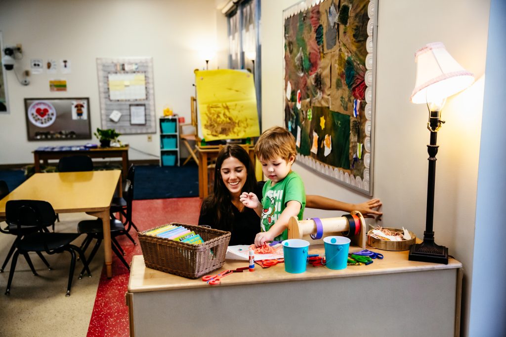 An early childhood teacher sits at a desk with a young child while working on a craft.
