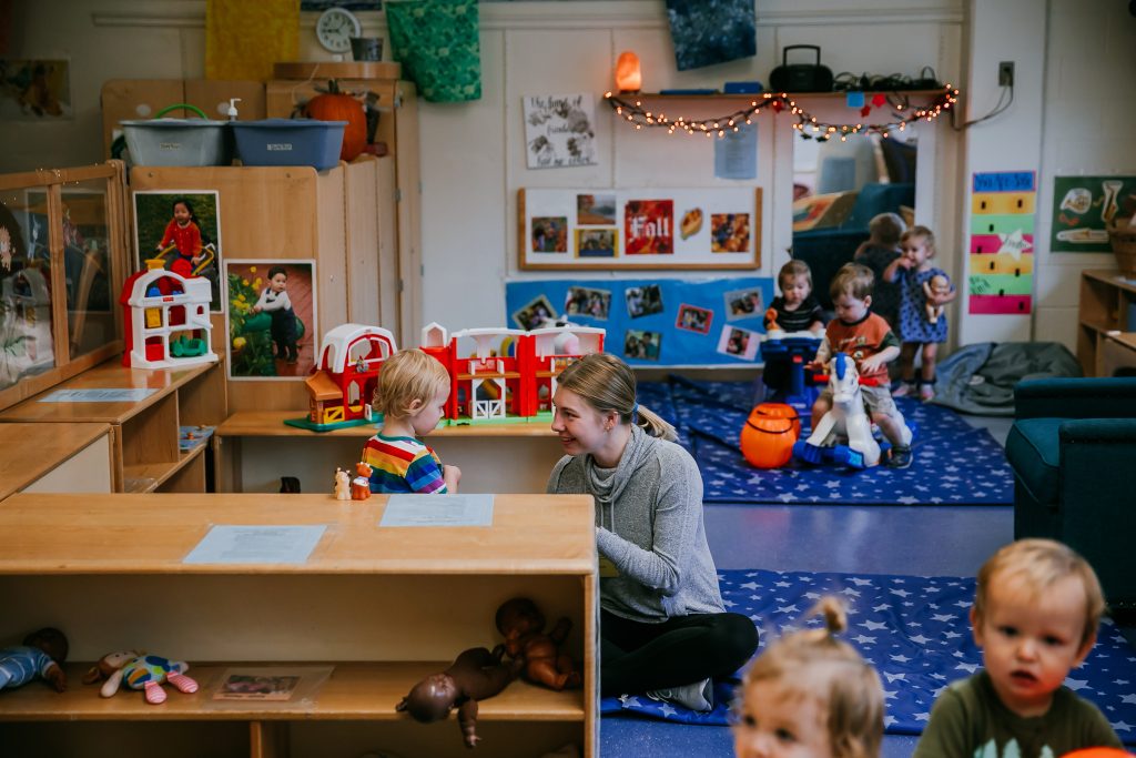 An early childhood educator plays with a a young child while others play at an early childhood education center.